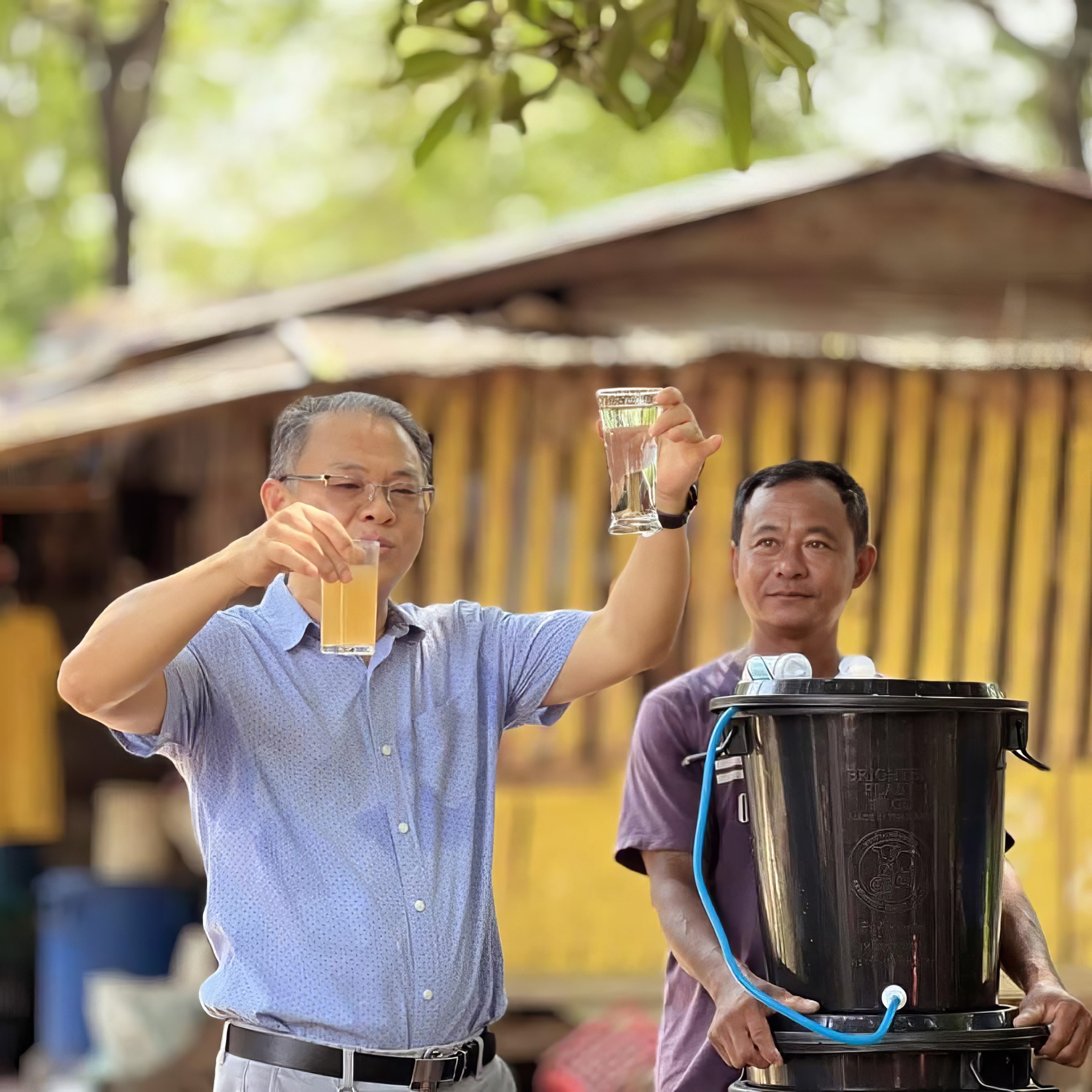 A man holds up a glass of filtered water beside a Filter of Hope bucket system